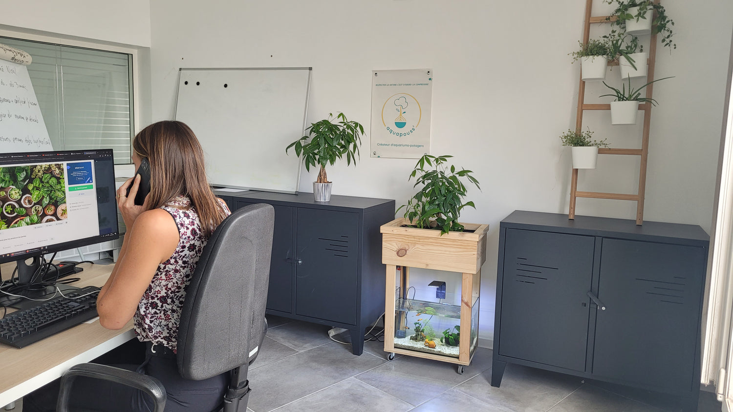 On peut voir une dame qui est assise à son bureau, au téléphone devant son ordinateur.A côté de son bureau entre deux étagères se trouve un kit aquapouss' mini, on peut voir l'aquarium bien éclairé et des plantes vertes dans le bas de culture. Il y a en plus quelques plantes en plus dans la pièce se qui donne une ambiance zen